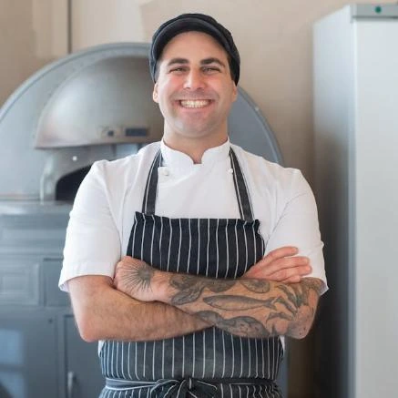 Homem sorridente de braços cruzados usando uniforme de padeiro com camisa branca, avental listrado e boina preta. Ele tem tatuagens visíveis nos braços e está em frente a um forno industrial, em um ambiente que aparenta ser uma padaria ou cozinha profissional.